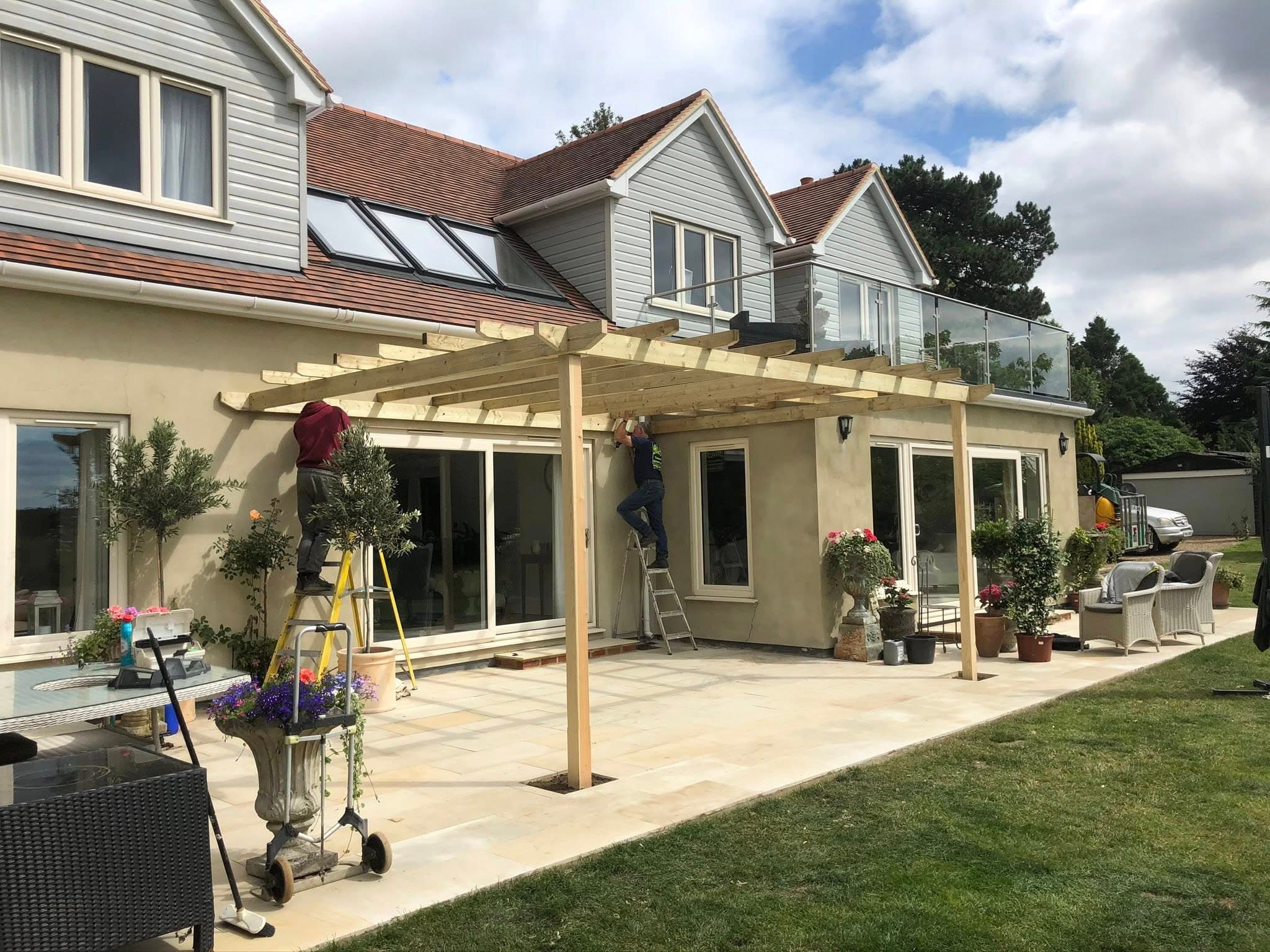 The M&M team installing a glass-balustrade pergola at a Kent country property — fresh sandstone patio, finished landscaping and a team member on the ladder fitting the canopy.